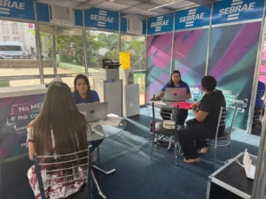 Fotografia colorida do interior de uma estrutura móvel de atendimento do Sebrae, montada em uma praça pública. No espaço climatizado, há duas mesas de vidro com computadores portáteis onde duas funcionárias do Sebrae, vestindo camisetas azuis da instituição, realizam atendimento presencial a duas mulheres sentadas em cadeiras de metal.
