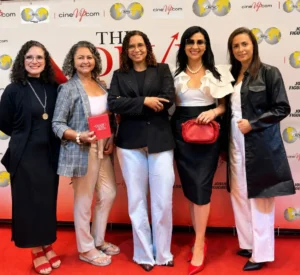 Cinco mulheres sorridentes, associadas da BPW Rondonópolis, posam lado a lado em um tapete vermelho durante um evento de cinema. Ao fundo, há um painel branco com as logomarcas da BPW e do portal CineVip.com. Da esquerda para a direita, a primeira mulher usa óculos, vestido e casaco longos pretos e sandálias vermelhas; a segunda veste blazer xadrez cinza, calça clara e segura a caixa escrito "O Diabo Veste Prada" na cor vermelha; a terceira usa blazer preto e calça jeans clara; a quarta veste uma blusa branca com babados, saia preta, óculos escuros e carrega uma bolsa vermelha; a quinta veste um casaco longo preto sobre blusa e calça brancas.