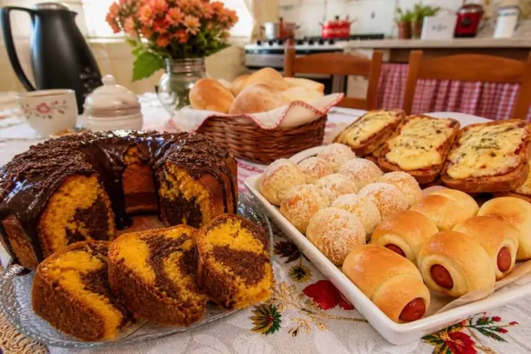 Lanche da tarde fácil com bolo de cenoura mesclado, salgadinho de salsicha, nozinho doce e torrada sabor pizza sobre mesa servida.