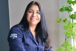 Mulher de cabelos longos e pretos, sorrindo gentilmente para a câmera. Ela veste um jaleco azul-marinho com o logotipo "Membro Oficial CITRG" visível na manga. O fundo é composto por uma parede clara e uma planta verde à direita, transmitindo um ambiente profissional e acolhedor.