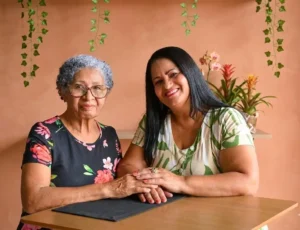 Duas mulheres, uma idosa com cabelos grisalhos e óculos e outra mais jovem com longos cabelos pretos, estão sentadas lado a lado sorrindo. Elas estão com as mãos dadas sobre uma mesa. A idosa usa um vestido preto floral e a mais jovem um vestido branco com estampas de folhas verdes. O fundo é uma parede terracota com plantas e flores.