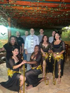 Fotografia de um grupo de dez pessoas em um restaurante com decoração rústica. Ao fundo, seis pessoas da equipe do restaurante posam para a foto, com o responsável pela cozinha ao centro usando um uniforme branco de chef. À frente, quatro bailarinas do grupo Handaras aparecem sorridentes, vestindo roupas pretas de dança do ventre com detalhes em moedas douradas e segurando bengalas douradas. O teto do local é de madeira e está coberto com folhas verdes.