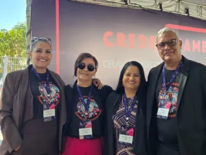 Quatro jornalistas da delegação de Rondonópolis — três mulheres e um homem — sorriem lado a lado posando para foto em Brasília. Eles estão em frente a um painel de credenciamento do Intercom com os logotipos do CEUB e da Intercom. Da esquerda para a direita: a primeira mulher usa blazer marrom; a segunda veste calça rosa e óculos escuros; a terceira veste uma blusa estampada com blazer escuro; e o homem, à direita, usa óculos de grau e blazer preto. Todos usam os crachás oficiais do evento pendurados no pescoço.