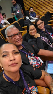Selfie vertical tirada em um auditório com poltronas escuras e detalhes em madeira. Em primeiro plano, quatro jornalistas de Rondonópolis aparecem sentados na mesma fileira, sorrindo para a foto. Na frente, à esquerda, uma mulher de cabelos presos e brincos de pérola segura a câmera. Atrás dela, está um homem de óculos e cabelos grisalhos. Ao lado dele, uma mulher de óculos sorri com a mão no queixo e, na ponta direita, outra mulher de óculos. Todos usam crachás oficiais do evento. Ao fundo, outras pessoas circulam pelas fileiras do auditório.