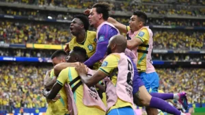 Fotografia colorida em plano médio capturada dentro de um estádio de futebol lotado. A imagem mostra a celebração intensa e calorosa de jogadores da seleção brasileira após a vitória. No centro, um grupo de atletas se abraça fortemente, expressando grande alívio e euforia. Entre eles, Vinícius Júnior aparece com a boca bem aberta em um grito de comemoração, vestindo a tradicional camisa amarela da seleção. Outros jogadores ao redor usam coletes de treino rosa claro sobre o uniforme e shorts azuis, enquanto o goleiro, vestindo uma camisa roxa, é erguido no ar durante o abraço coletivo. Ao fundo, as arquibancadas do estádio aparecem desfocadas, completamente tomadas por uma multidão de torcedores vestindo as cores verde e amarela.