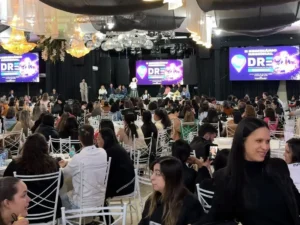 Fotografia colorida em plano aberto mostrando o interior do Espaço Tulipas durante um evento educacional. O salão está completamente preenchido por um grande público sentado em mesas redondas com toalhas claras e cadeiras brancas de ferro, a maioria de costas ou de perfil para a câmera. Ao fundo, há um palco elevado com cortinas pretas onde várias autoridades estão sentadas em linha, e uma mulher de blusa clara e saia escura está de pé no centro falando ao microfone. Acima do palco, destacam-se globos espelhados de discoteca pendurados e grandes lustres de cristal acesos. Três grandes painéis de LED exibem a identidade visual roxa do evento com a inscrição "II SEMINÁRIO REGIONAL DRE". Em primeiro plano, no canto inferior direito, destaca-se o rosto de perfil de uma mulher sorrindo, de cabelos pretos e lisos, vestindo blusa escura. A iluminação do ambiente é interna e cênica.