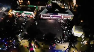 Fotografia aérea noturna, tirada de um ângulo superior (vista de drone), mostrando o pátio da Festa das Nações em Rondonópolis. No espaço iluminado, é possível ver diversas barracas cobertas e enfileiradas que representam as culinárias dos países, com voluntários e visitantes se movimentando entre elas. Há mesas distribuídas pela área externa e filas organizadas de pessoas aguardando atendimento em frente aos estandes gastronômicos. Árvores e coqueiros aparecem integrados ao cenário, dividindo os setores da feira beneficente.