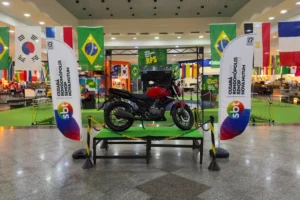Fotografia em plano médio do interior do Rondon Plaza Shopping, mostrando o espaço decorado para a campanha. No centro, sobre um tablado baixo com grama artificial verde e cercado por correntes de sinalização amarelas e pretas, está em exibição uma motocicleta de modelo urbano na cor vermelha e preta. Nas laterais do tablado, há dois tótens verticais brancos com o logotipo colorido do SBT e o texto "Cuiabá, Rondonópolis, Sinop, Nova Mutum". Ao fundo, a decoração inclui bandeiras de diversos países penduradas no teto, com destaque para a bandeira do Brasil, além de uma estrutura metálica com uma placa verde onde se lê "Entre em campo com o RPS". O piso de granito cinza reflete as luzes do teto do shopping.