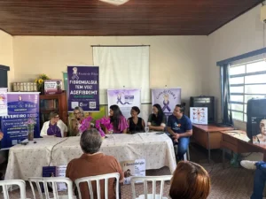 Fotografia colorida em plano médio de uma reunião em uma sala com teto de madeira escura. Atrás de uma mesa comprida, coberta com uma toalha clara estampada com flores, estão sentadas seis pessoas alinhadas. Da esquerda para a direita: uma mulher de blazer branco e blusa roxa; uma mulher de óculos e blusa branca estampada que segura um microfone e fala; uma mulher de blusa rosa escuro; uma mulher de blusa preta regata; uma mulher de blusa verde escura e óculos; e um homem de camiseta azul-marinho com um logotipo redondo no peito. No centro da mesa, há vasos com orquídeas em tons de rosa e roxo. Ao fundo, fixados na parede branca, destacam-se banners roxos da Associação Gente de Fibra com mensagens como "FIBROMIALGIA TEM VOZ!". Em primeiro plano, aparecem as costas de duas pessoas na plateia assistindo ao evento: um homem com camisa marrom à esquerda e uma mulher à direita.