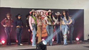 Fotografia colorida mostrando um grupo de oito mulheres alinhadas lado a lado em um palco de desfile, vestindo trajes de estilo country. Elas usam chapéus de caubói em tons de marrom, preto, rosa e branco, camisas de manga longa (nas cores vinho, azul-marinho, rosa, bege e branca) e calças jeans justas com cintos de fivelas grandes. Três das mulheres posicionadas ao centro usam faixas de premiação transversais no peito, onde se leem os títulos "Garota Rodeio", "Garota Exposul" e "Garota Country". Ao fundo, há um painel escuro com uma estrutura circular iluminada no centro e refletores de luz cênica avermelhada nas laterais do palco. Na parte inferior da imagem, aparece o topo de um refletor de luz branco focado para cima.