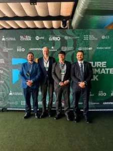 Fotografia em plano inteiro de quatro homens vestidos formalmente com ternos e crachás do evento, posados lado a lado para a foto. Eles estão em pé sobre um tapete verde que imita grama, diante de um grande painel verde-escuro com logotipos de marcas parceiras e a inscrição parcial "Nature & Climate Week". O primeiro homem à esquerda veste terno azul-claro; o segundo veste terno escuro com camisa branca aberta; o terceiro veste terno cinza; e o quarto, posicionado à direita, veste terno preto com gravata azul texturizada. O piso reflete sutilmente a iluminação do ambiente.