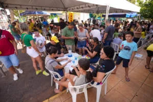 Fotografia colorida e ampla de uma movimentada ação comunitária realizada ao ar livre, embaixo de grandes tendas brancas. Em primeiro plano, adultos e crianças estão sentados ao redor de mesas de plástico brancas; algumas pessoas preenchem papéis e formulários, enquanto outras conversam ou usam o celular. No centro, uma menina de perfil segura um papel branco e conversa com uma mulher. Ao redor e ao fundo, dezenas de pessoas circulam pelo espaço, várias delas vestindo camisas verdes e amarelas da seleção brasileira. Uma bandeira do Brasil é visível ao longe, no canto superior esquerdo, entre árvores e outras tendas. O chão é dividido entre o asfalto escuro da rua e uma calçada de azulejo claro.
