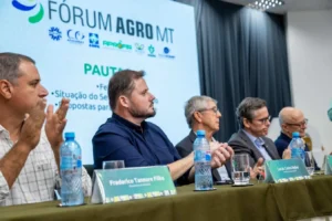 Uma fotografia em ângulo lateral médio de um painel de discussão em uma conferência. Cinco homens estão sentados em uma mesa longa coberta com uma toalha verde-oliva. Eles estão voltados para a direita, ouvindo um orador (fora do enquadramento). Identificações de mesa verdes estão na frente de cada homem. O homem mais próximo à esquerda tem uma identificação legível que diz: "Frederico Tannure Filho" e "Presidente da Famato". Ele está sentado e ouvindo. O homem ao centro está gesticulando levemente com as mãos enquanto fala ou concorda. Outras garrafas de água e copos estão espalhados pela mesa. No fundo, há um grande ecrã de projeção branco que exibe o logotipo "FÓRUM AGRO MT" em letras grandes e coloridas, com logotipos menores abaixo e uma lista de pauta que começa com "PAUTA". Uma cortina de palco azul escura está ao lado do ecrã. A iluminação é de conferência, uniforme e clara.