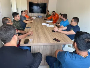 Uma foto em plano médio mostra uma reunião do Comitê Municipal de Gestão do Fogo dentro de uma sala. Doze pessoas estão sentadas ao redor de uma grande mesa retangular de madeira clara. Entre os participantes, do lado esquerdo, destaca-se um homem com fardamento camuflado do Exército; ao fundo, um homem veste o uniforme laranja do Corpo de Bombeiros; e no canto inferior direito, um homem indígena com colar tradicional participa da discussão. Na parede ao fundo, há um monitor de TV desligado. O clima é de debate e atenção.
