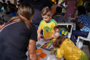 Foto em plano médio e ângulo levemente superior mostrando crianças concentradas ao redor de uma mesa branca de plástico ao ar livre. No centro, um menino em pé veste uma camiseta amarela do Brasil e segura figurinhas nas mãos, olhando para baixo. Sentada ao seu lado direito, uma menina de cabelos castanhos, usando uma camiseta amarela e um grande laço verde e amarelo na cabeça, observa a movimentação. Sobre a mesa, há um álbum de figurinhas ilustrado da Copa do Mundo, bloquinhos de figurinhas repetidas e pequenos organizadores plásticos cor-de-laranja. Adultos e outras pessoas ao redor aparecem parcialmente na imagem, criando um ambiente movimentado e familiar.