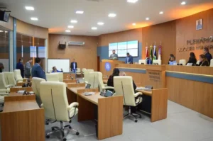 A imagem mostra o interior do Plenário Ulysses Guimarães da Câmara Municipal de Rondonópolis, conforme indicado pelo letreiro na parede de madeira. O ambiente é amplo, com painéis de madeira cobrindo as paredes e o teto, que possui iluminação embutida. No centro do plenário, há várias bancadas de madeira individuais com cadeiras de couro bege, dispostas em fileiras, de frente para a mesa diretora. Na mesa diretora, ao fundo, estão posicionadas três pessoas sentadas: um homem de terno azul escuro e duas mulheres de roupa escura. Atrás deles, na parede de madeira, estão penduradas as bandeiras do Brasil, do estado de Mato Grosso e do município de Rondonópolis. Um grande ecrã de projeção está localizado na parede ao lado da mesa diretora. No centro do plenário, em pé e de costas para a câmera, estão dois homens de terno azul, observando a mesa diretora. Uma pessoa está sentada em uma das bancadas centrais. Na parede ao lado esquerdo, há grandes janelas de vidro que dão para uma área externa. O teto possui iluminação embutida e um aparelho de ar-condicionado está visível na parede esquerda.