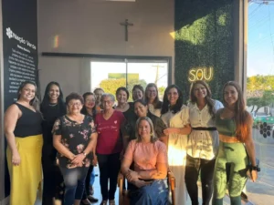 Fotografia em ambiente interno mostrando um grupo de treze mulheres sorridentes reunidas em uma loja. Uma mulher de camiseta rosa claro e saia jeans está sentada em uma cadeira de madeira no centro, enquanto as outras doze mulheres estão de pé ao redor dela, vestindo roupas casuais variadas (como blusas floridas, vestidos brancos, roupas pretas e um conjunto de academia verde). Ao fundo, há uma parede cinza com um crucifixo acima de uma porta de vidro, um painel preto à esquerda com o texto "Nação Verde - Nossa História" escrito em branco, e à direita uma parede de folhagem verde com um letreiro luminoso amarelo que diz "SOU". A luz do sol entra pela janela à direita, iluminando suavemente o grupo.
