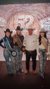 otografia colorida mostra quatro pessoas em pé no centro de um palco com fundo iluminado. Da esquerda para a direita, uma jovem com faixa azul de "GAROTA COUNTRY", a vencedora no centro com faixa dourada de "GAROTA EXPOSUL", um homem de camisa branca e calça jeans, e uma jovem com faixa de "GAROTA RODEIO". Todos usam chapéus de cowboy e sorriem.