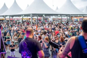 Uma fotografia em ângulo médio e plano aberto, tirada a partir do palco, mostrando a visão dos músicos para o público de um festival ao ar livre. Em primeiro plano, de costas para a câmera, aparecem partes dos corpos de dois integrantes de uma banda, com destaque para as costas de um homem vestindo camiseta preta à esquerda e outro com regata preta e alça de instrumento à direita. Diante deles, uma grande multidão de pessoas de diversas idades assiste ao show em pé. No público, destacam-se uma jovem de cabelos ruivos e saia xadrez tirando uma foto com o celular, uma criança com camiseta preta à esquerda e várias outras pessoas prestando atenção ao palco. Ao fundo, há uma grande estrutura de tendas brancas com coberturas em formato de pirâmide pontiaguda, abrigando diversas barracas de alimentação do evento. A iluminação é natural, típica de um dia nublado de fim de tarde.