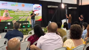 Uma fotografia panorâmica em plano médio mostra uma palestra técnica ocorrendo dentro de um pavilhão da Exposul. No palco elevado, um palestrante masculino de camisa polo verde e calça jeans fala ao microfone e gesticula com a mão esquerda. Atrás dele, um grande painel de LED exibe o slide da apresentação com o título "Crédito Rural e Regularização Ambiental e Fundiária: Como isso impacta o seu negócio", acompanhado da marca VÉRTICE e de um gráfico de um saco de dinheiro com cifrão sobre um campo agrícola. Diante do palco, uma plateia atenta composta por produtores rurais e membros da comunidade ocupa várias fileiras de cadeiras plásticas brancas, vistos de costas. À direita, dois profissionais de captação de imagem, um cinegrafista e um fotógrafo, registram o evento com equipamentos profissionais e iluminação de estúdio. O ambiente é amplo, com cortinas pretas ao fundo e sistemas de ar condicionado visíveis.