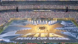 Uma fotografia panorâmica em plano geral e vista aérea captura a grandiosidade de um estádio lotado durante a cerimônia de encerramento da Copa do Mundo FIFA 2026. O gramado está transformado por uma megaestrutura de tecidos e luzes que forma um palco complexo e vibrante. No centro geométrico, uma representação luminosa e dourada do troféu da Copa do Mundo brilha intensamente, com raios de luz e estruturas arquitetônicas estendendo-se dele para fora, criando um padrão dinâmico. Em primeiro plano, estendido monumentalmente sobre o campo, um enorme banner traz o texto "FIFA WORLD CUP 2026" em grandes letras douradas e pretas. Fogos de artifício ou jatos de luz acendem no fundo do campo, próximo à área do palco principal. Milhares de espectadores preenchem as arquibancadas em camadas, visíveis ao redor de todo o estádio, criando uma atmosfera vibrante e lotada sob a luz do dia.