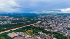 Fotografia aérea panorâmica em plano geral mostrando a cidade de Rondonópolis recortada pelo Rio Vermelho. O rio, de águas barrentas e tom marrom, serpenteia pelo centro da imagem, ladeado por extensas faixas de mata ciliar verde e densa. À direita e ao fundo, a área urbana se espalha com uma mistura de casas de telhado cerâmico, ruas desenhadas e edifícios verticais concentrados. Na parte inferior esquerda, destaca-se a estrutura de um grande centro comercial e seu estacionamento. O horizonte ao fundo revela áreas rurais e de vegetação sob um céu azul coberto por nuvens brancas e carregadas.