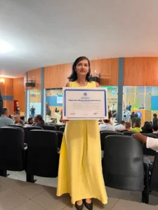 Uma fotografia em close médio de uma mulher de pé em um auditório em Rondonópolis, no Mato Grosso. Ela tem cabelos escuros na altura dos ombros e veste um vestido amarelo longo com um colar de pérolas. Ela segura, com as duas mãos, um grande certificado emoldurado com a inscrição "Título de Cidadã Rondonopolitana" em letras grandes e azuis. O documento está assinado por várias autoridades municipais. A mulher sorri para a câmera. Ao fundo, um auditório com assentos pretos está repleto de pessoas sentadas. Atrás delas, há painéis de madeira e vidro. Do outro lado do vidro, uma sala de reuniões oficial com pessoas sentadas a uma mesa grande é visível. O brasão do município de Rondonópolis está na parede ao fundo. A iluminação ambiente é quente, proveniente de luminárias embutidas no teto.
