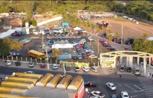 Uma vista aérea capturada por drone mostra um grande evento automotivo ao ar livre no Parque das Águas, em Rondonópolis, Mato Grosso, realizado durante o pôr do sol. Centenas de carros clássicos de várias cores estão alinhados em estacionamentos e áreas de gramado. Perto da entrada principal com arco, um ônibus escolar amarelo brilhante está estacionado. Barracas brancas e tendas de expositores estão montadas no centro do evento, com palmeiras e áreas verdes circundantes.