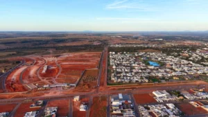 Uma fotografia aérea panorâmica em plano geral capturada no final da tarde, mostrando a infraestrutura viária e o desenvolvimento urbano em Rondonópolis, Mato Grosso. Uma longa rodovia corta verticalmente o centro da imagem, dividindo a paisagem. Do lado direito, há um bairro residencial planejado com casas de cor clara e um pequeno lago azul cercado por árvores. Do lado esquerdo, extensas áreas de terra vermelha preparada indicam novos loteamentos em construção. Na parte inferior, vias urbanas e edificações comerciais completam o cenário. O horizonte distante apresenta colinas suaves sob um céu claro que transita de tons de azul a um dourado suave devido ao pôr do sol.