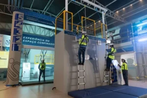 Fotografia tirada no interior de um pavilhão industrial moderno e amplo, capturando um treinamento prático de segurança no Sesi Experience. A estrutura central é um grande simulador de contêiner e plataforma de concreto cinza. Nele, dois trabalhadores, vestindo uniformes profissionais, capacetes brancos, luvas e cintos de segurança completos acoplados a um sistema de cabos de aço e roldanas aéreas, sobem duas escadas de alumínio paralelas. Um deles está mais alto, quase no topo da plataforma protegida por guarda-corpos amarelos. No topo da plataforma, há outra pessoa observando o procedimento. À esquerda, ao nível do chão, um instrutor, também de capacete e cinto de segurança, supervisiona a operação próximo a um painel de controle branco. Ao fundo, um grande painel de LED exibe as marcas do Sistema Fiemt, Sesi e Senai, e o texto 'SESI EXPERIENCE' e 'TREINAMENTO EM ALTURA'. À direita, outro profissional aguarda a sua vez. A iluminação é focada e eficiente.