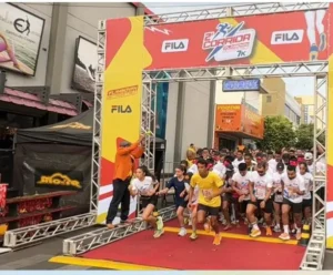 Uma fotografia colorida captura o momento exato da largada de uma corrida de rua. Dezenas de corredores, a maioria usando camisetas brancas e números de peito, cruzam uma linha de partida no asfalto. Eles correm em direção à câmera, sob uma grande estrutura de pórtico de metal inflável.