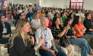 Uma vista angular de uma plateia em um auditório, focada nos espectadores sentados nas primeiras fileiras. Muitas pessoas estão voltadas para a esquerda, olhando para algo fora do quadro. Em primeiro plano, uma mulher loira com blusa preta sorri enquanto segura um smartphone, e ao lado dela, um homem com uma camisa social clara e crachá de pescoço segura um celular. Mais à direita, uma mulher oriental de bolinhas tira uma foto com outro celular. A sala está cheia e bem iluminada.