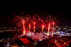 Vista aérea noturna do Parque de Exposições de Rondonópolis durante a 52ª Exposul. A imagem mostra uma multidão aglomerada em frente ao palco principal, que está iluminado. Várias rajadas de fogos de artifício vermelhos e laranjas explodem no céu escuro acima da arena, criando um grande dossel de luz. A cidade ao fundo está visível com luzes dispersas. (139 caracteres)