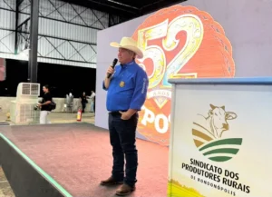 Fotografia de um homem de meia-idade, vestindo chapéu de cowboy branco, camisa social azul e calça jeans, falando ao microfone. Ele está em um palco, de pé sobre um tablado com carpete vermelho, posicionado em frente a um grande painel decorativo com o número "52" estilizado e a inscrição "EXPOSUL". À direita, há um púlpito branco com o logotipo do "Sindicato dos Produtores Rurais de Rondonópolis". Ao fundo, um galpão industrial e algumas pessoas desfocadas.