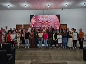 Foto panorâmica do Auditório Professora Coraci, no Núcleo de Rondonópolis da UNEMAT, mostrando um grupo de dezenas de pessoas sorrindo e em pé no palco durante o lançamento oficial do Instituto Consciência Curativa (ICC). No fundo, ao centro, um grande telão de LED exibe o logotipo dourado da flor de lótus e o nome "Instituto Consciência Curativa" em letras brancas sobre fundo rosa, ladeado por projeções de flores. A plateia está vazia em primeiro plano.