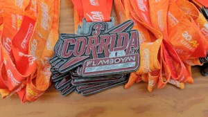 Uma imagem de close-up mostrando um conjunto de medalhas de participação da "3ª Corrida de Rua Flamboyan Esportes". As medalhas são de cor prateada com detalhes em relevo nas cores vermelha e preta, com o texto "CORRIDA DE RUA 7K FLAMBOYAN Esportes". Elas estão penduradas em fitas laranja vibrante que trazem estampas brancas e vermelhas com os textos "30 de Agosto Rondonópolis - MT". As medalhas estão apoiadas em uma superfície de madeira.