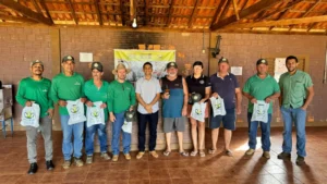 Fotografia em ambiente coberto com parede de tijolos à mostra e estrutura de teto em madeira. Ao centro, um grupo de dez pessoas está posicionado lado a lado para uma foto institucional. A maioria veste camisetas verdes ou camisas de manga curta, bonés e segura sacolas brancas com o logotipo da Aprosmat impresso em verde e amarelo. Ao fundo, no centro da parede, há um banner do programa "Motorista Nota 10" sobre o carregamento e transporte de sementes.