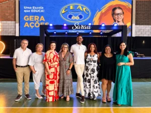 Um grupo de oito pessoas, sendo um homem e sete mulheres, posa sorrindo para uma foto em pé em um ginásio. Eles estão em frente a uma estrutura de metal preta e a um grande painel azul com o logotipo do CEAC Sistema Positivo, o slogan "Uma Escola que EDUCA! GERA AÇÕES" e a imagem de uma menina de óculos. O homem está no centro do grupo, vestindo uma camisa polo branca e calça bege. As mulheres usam roupas casuais e coloridas, algumas estampadas. O chão do ginásio é verde com linhas amarelas e brancas. Há iluminação cênica na estrutura preta.