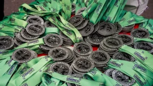 Uma fotografia em close de centenas de medalhas circulares de metal escura e prata, empilhadas em uma mesa. Cada medalha tem um design em relevo e está presa a uma fita de cetim verde-clara com o logotipo "UNIMED" repetido. O texto "CORRIDA UNIMED RONDONÓPOLIS" e "2025" é visível em várias medalhas e fitas. Uma única medalha dourada está ligeiramente deslocada à direita do centro. No canto superior direito, vê-se a parte inferior de uma perna e pé humano.