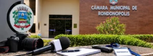 Fotografia de equipamentos de imprensa, como câmera profissional, microfone, gravador, bloco de notas e crachá escrito "Imprensa", dispostos sobre um balcão de granito, com o prédio da Câmara Municipal de Rondonópolis ao fundo.