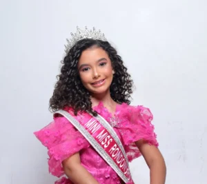 Um retrato de estúdio em close-up de uma jovem, Helena Leão, Mini Miss Rondonópolis 2026. Ela está posicionada no centro e ligeiramente à direita da moldura, sorrindo diretamente para a câmera com uma expressão alegre e confiante. Ela tem cabelos castanhos escuros cacheados e volumosos, que estão arrumados e fluindo sobre os ombros. Ela usa uma coroa prateada cravejada de diamantes no topo da cabeça. Ela veste um vestido de gala rosa brilhante com apliques de flores 3D e detalhes de strass. Sobre o ombro direito e cruzando o busto está uma faixa oficial branca com bordas em fita de cetim rosa e letras prateadas, onde se lê "MINI MISS RONDONÓPOLIS" e mais abaixo, em letras menores, "MATO GROSSO". Ela também usa brincos de argola pequenos e maquiagem leve. O fundo é uma parede de estúdio branca simples, sem distrações. A iluminação é suave e uniforme.