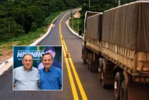 Fotografia de uma rodovia recém-pavimentada e sinalizada com faixas amarelas no centro, cercada por vegetação. À direita, um caminhão de carga de grande porte trafega pela pista de asfalto. No canto inferior esquerdo, há um quadro em destaque com a imagem de dois homens sorrindo lado a lado em um evento institucional.