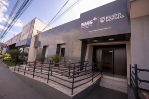 Fotografia em ângulo externo da fachada do prédio da Secretaria Municipal de Saúde (SMS) em Rondonópolis. A estrutura possui fachada moderna em tom cinza-escuro com espelhamento, onde se lê a sigla "SMS - Secretaria Municipal de Saúde" e o brasão do município. Na entrada, há uma rampa de acessibilidade com corrimão de metal preto, degraus e pequenas plantas ornamentais no canteiro ao lado.