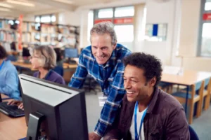 Dois homens sorridentes em um ambiente de estudo e informática. O homem em primeiro plano tem cabelos cacheados escuros, veste jaqueta marrom e está sentado em frente a um monitor de computador. Ao seu lado, em pé e inclinado para ver a tela, um homem mais velho de cabelos grisalhos e camisa xadrez azul sorri enquanto observa o monitor. Ao fundo, outras pessoas trabalham em computadores em um ambiente iluminado por janelas.