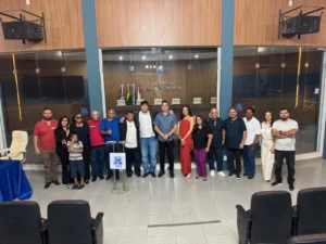 Fotografia colorida tirada de um ângulo elevado, mostrando um grupo de dezoito pessoas (dezessete adultos e uma criança) posando para foto no plenário Ulysses Guimarães da Câmara Municipal de Rondonópolis. Eles estão de pé, alinhados lado a lado no centro do plenário, em frente a uma divisória de vidro transparente que revela ao fundo a mesa diretora e as cadeiras dos vereadores. À esquerda, vê-se parte de uma mesa coberta com toalha azul. No centro, à frente do grupo, há um púlpito de metal com o brasão do município em azul e branco. As pessoas vestem roupas casuais e sociais variadas e sorriem para a câmera. O ambiente é amplo, com piso de lajotas claras e paredes com detalhes em madeira. À frente, no primeiro plano inferior, vê-se o encosto de duas fileiras de cadeiras escuras do auditório. A iluminação é artificial e uniforme.