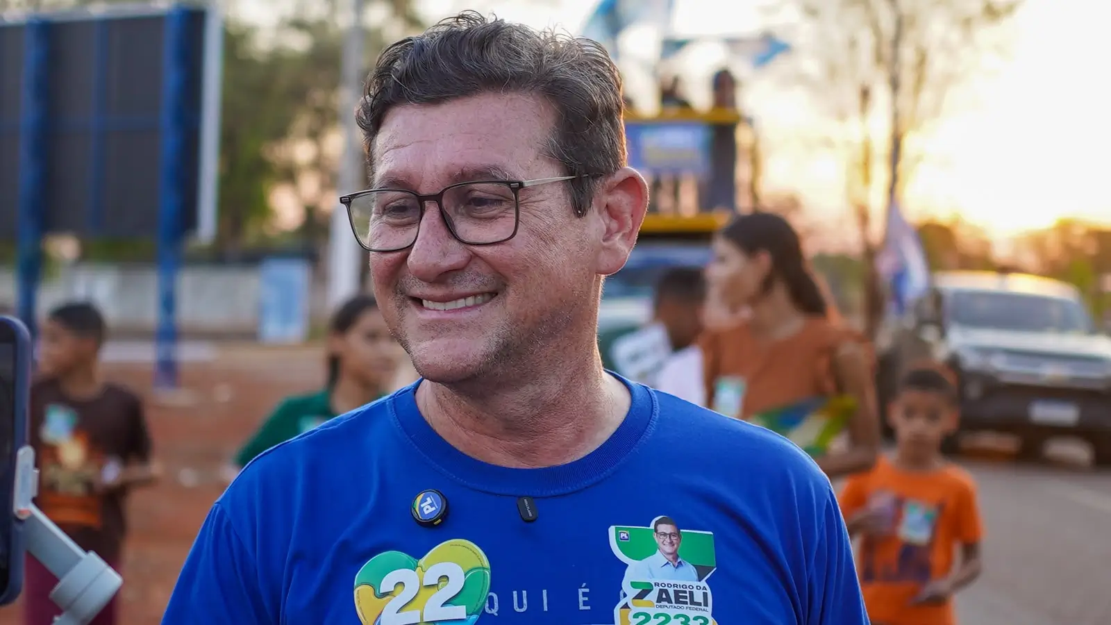 Uma fotografia colorida mostra Rodrigo da Zaeli, um homem de meia-idade, com cabelos castanhos curtos e grisalhos e óculos de grau, sorrindo e olhando para o lado esquerdo. Ele veste uma camiseta azul royal com broches de campanha: um redondo com o número "22" nas cores do Brasil (amarelo, verde e azul), outro do "PL", e um quadrado com sua foto e o nome "RODRIGO DA ZAELI DEPUTADO FEDERAL 2233". O fundo está desfocado, indicando um evento de rua ao ar livre, com outras pessoas e um trio elétrico borrados ao longe durante o dia.
