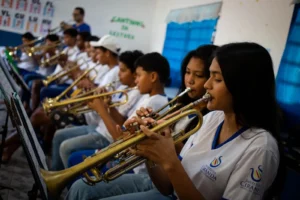Esta é uma foto horizontal de profundidade média em uma sala de aula de música, mostrando duas jovens garotas e outros alunos no fundo, todos tocando trompetes. A jovem garota na frente tem longos cabelos escuros e segura o trompete dourado com as duas mãos, perto de seu lábio, olhando para a partitura à sua esquerda. Ela usa uma camiseta branca com uma linha azul ao redor do pescoço e duas logomarcas que dizem "INSTITUTO CIRANDA" e "MÚSICA E CIDADANIA". Outra jovem garota ao seu lado também está tocando o instrumento. Ambas vestem a mesma camiseta branca. O garoto atrás delas está segurando o trompete e olhando para a frente. O resto dos alunos em uma fileira está focado em seus próprios instrumentos dourados. Eles estão sentados em cadeiras de plástico azul. Há paredes brancas e uma cortina azul na parede atrás. A logomarca do Instituto Ciranda, Música e Cidadania, está impressa no lado direito e no centro da camiseta de ambas as garotas da frente.