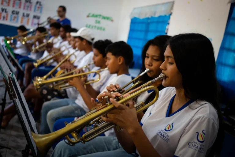 Esta é uma foto horizontal de profundidade média em uma sala de aula de música, mostrando duas jovens garotas e outros alunos no fundo, todos tocando trompetes. A jovem garota na frente tem longos cabelos escuros e segura o trompete dourado com as duas mãos, perto de seu lábio, olhando para a partitura à sua esquerda. Ela usa uma camiseta branca com uma linha azul ao redor do pescoço e duas logomarcas que dizem "INSTITUTO CIRANDA" e "MÚSICA E CIDADANIA". Outra jovem garota ao seu lado também está tocando o instrumento. Ambas vestem a mesma camiseta branca. O garoto atrás delas está segurando o trompete e olhando para a frente. O resto dos alunos em uma fileira está focado em seus próprios instrumentos dourados. Eles estão sentados em cadeiras de plástico azul. Há paredes brancas e uma cortina azul na parede atrás. A logomarca do Instituto Ciranda, Música e Cidadania, está impressa no lado direito e no centro da camiseta de ambas as garotas da frente.