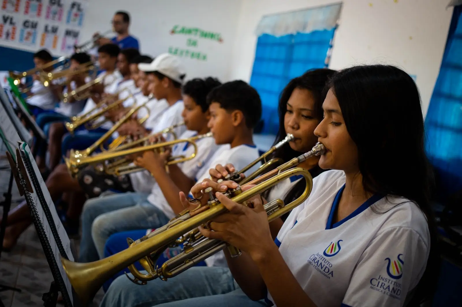 Esta é uma foto horizontal de profundidade média em uma sala de aula de música, mostrando duas jovens garotas e outros alunos no fundo, todos tocando trompetes. A jovem garota na frente tem longos cabelos escuros e segura o trompete dourado com as duas mãos, perto de seu lábio, olhando para a partitura à sua esquerda. Ela usa uma camiseta branca com uma linha azul ao redor do pescoço e duas logomarcas que dizem "INSTITUTO CIRANDA" e "MÚSICA E CIDADANIA". Outra jovem garota ao seu lado também está tocando o instrumento. Ambas vestem a mesma camiseta branca. O garoto atrás delas está segurando o trompete e olhando para a frente. O resto dos alunos em uma fileira está focado em seus próprios instrumentos dourados. Eles estão sentados em cadeiras de plástico azul. Há paredes brancas e uma cortina azul na parede atrás. A logomarca do Instituto Ciranda, Música e Cidadania, está impressa no lado direito e no centro da camiseta de ambas as garotas da frente.