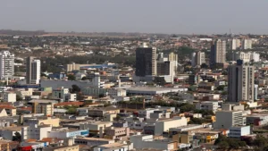 Fotografia panorâmica e aérea da área urbana de Rondonópolis, capturada de um ponto elevado durante o dia sob céu claro. A imagem destaca a densidade de prédios comerciais e residenciais, além de casas e galpões comerciais espalhados pelo centro e pelos bairros. Em primeiro plano e no centro, destacam-se diversos edifícios de portes variado, entre prédios mais altos com fachadas espelhadas e claras e construções térreas de comércio e residências. Ao fundo, avista-se a vegetação e o horizonte levemente ensolarado que delimita a extensão da cidade.